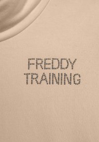 T-shirt beige con una texture liscia, caratterizzato dalla scritta "FREDDY TRAINING" in strass argentati, posizionata centralmente sul petto.