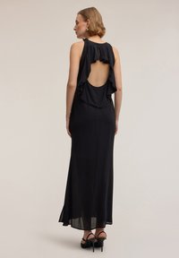 Abito maxi nero con apertura sulla schiena, volant sovrapposti sulle spalle e bordi decorati. Tessuto liscio con silhouette fluente e spacco alto.