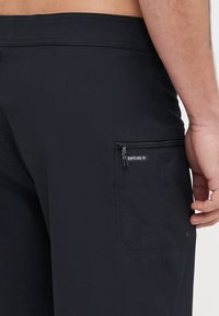 Schwarze Boardshorts mit einer Reißverschlusstasche und einem Logosticker, die über eine glatte Textur und eine taillierte Passform verfügen.