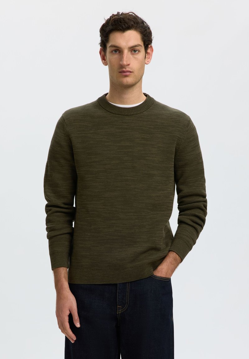 Homme aux cheveux foncés portant un pull en tricot vert olive et un jean bleu foncé, debout avec une main dans la poche devant un fond uni.