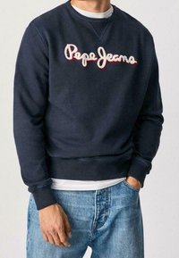 Hombre vistiendo un sudadera azul marino de Pepe Jeans con un logotipo blanco y rojo, camiseta blanca debajo y jeans azules, con una mano en el bolsillo.