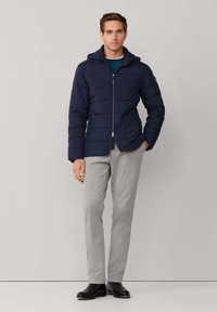 Chaqueta acolchada azul marino con cierre de cremallera, capucha y textura acolchada; combinada con pantalones gris claro y zapatos negros. Diseño minimalista.