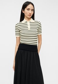 Ριγέ πλεκτό πόλο μπλουζάκι σε κρεμ με σκούρα πράσινες ρίγες, με γιακά και πλαίσιο με κουμπιά. Συνδυάζεται με μαύρη φούστα.