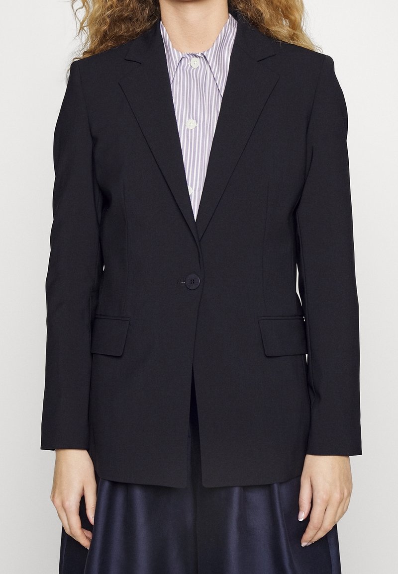 Femme portant un blazer noir à un bouton sur une chemise rayée violet clair et blanc, les mains détendues sur les côtés.
