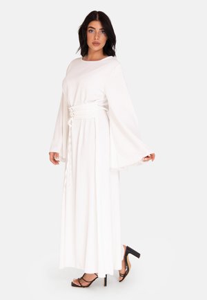 White maxi dress with long bell sleeves, a cinched waist with lace detail, made from a smooth fabric, paired with black strappy heels.