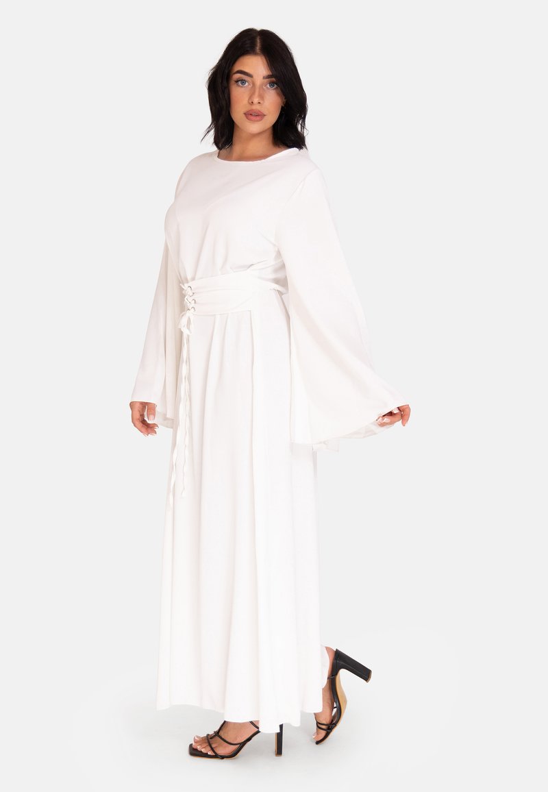 Une robe maxi blanche avec de longues manches en cloche, une taille cintrée avec des détails en dentelle, fabriquée à partir d'un tissu lisse, associée à des talons noirs à brides.