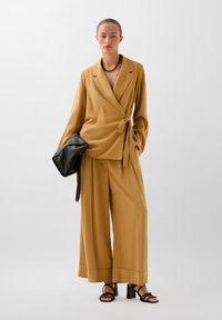 Senfgelb, wickelförmige Bluse kombiniert mit weit geschnittenen Hosen; glattes Material und strukturiertes Design. Eine schwarze Clutch und Riemchen-Absätze runden den Look ab.