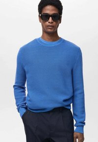 Blauw textuurtrui met een ronde hals en lange mouwen, gecombineerd met zwarte broeken. De stof heeft een geribd patroon.