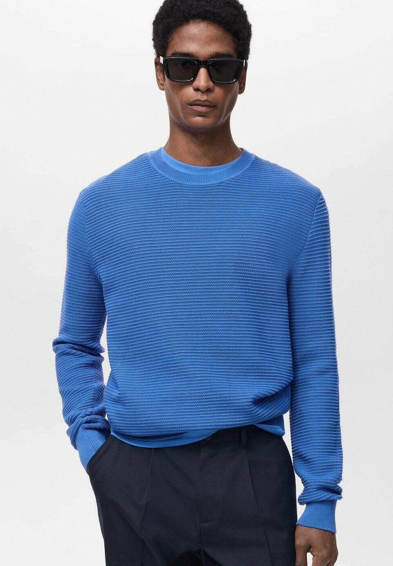 Blauw textuurtrui met een ronde hals en lange mouwen, gecombineerd met zwarte broeken. De stof heeft een geribd patroon.