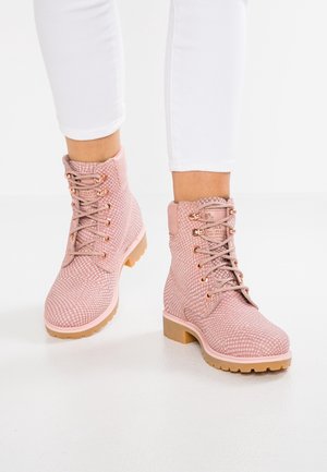 Osoba nosząca różowe, teksturowane, sznurowane botki za kostkę z beżowymi podeszwami oraz białe spodnie sięgające kostek na białym tle.