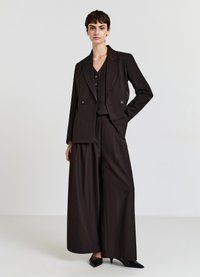 Costume sur mesure marron foncé comprenant un blazer croisé, un pantalon à jambes larges et un gilet ajusté avec fermeture à boutons. Escarpins noirs à bouts pointus.