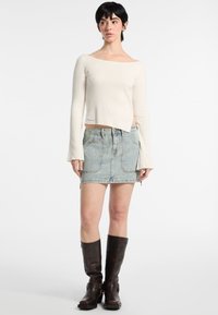 Maglione a spalle scoperte color crema a coste, mini gonna in denim azzurro con tasche frontali, stivali lunghi beige. I tessuti contrastano, abbigliamento casual.