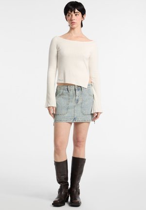 Pull en crème à épaules dénudées en maille côtelée, mini-jupe en denim bleu clair avec poches avant, bottes longues beiges. Contraste des textures, tenue décontractée.