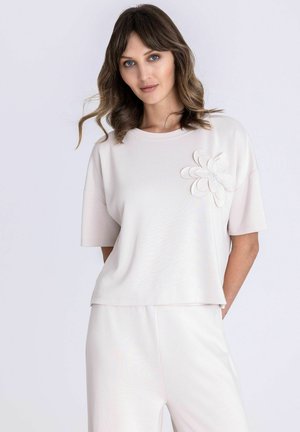 Frau mit welligem braunem Haar trägt ein lockeres blassrosa Kurzarmoberteil mit großem Blumendetail und passende Hose, stehend vor einfachem Hintergrund.