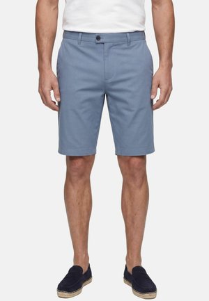Mann trägt hellblaue knielange Shorts, weißes Hemd in die Hose gesteckt und dunkelblaue Slip-On-Schuhe vor einfachem weißem Hintergrund.