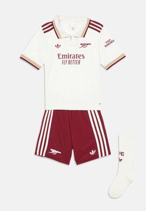 Uniforme juvenil del Arsenal FC que presenta una polo blanca de manga corta con detalles en carmesí, pantalones cortos carmesí a juego con franjas blancas y un calcetín blanco.