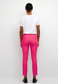 Pantalon rose ajusté avec une texture lisse, deux poches arrière, et une coupe fuselée, associé à un t-shirt blanc uni et des chaussures noires.
