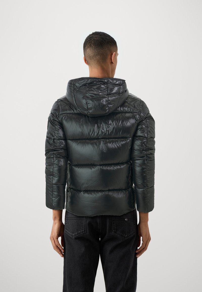 Giubbotto piumino verde scuro con cappuccio, finitura lucida, design trapuntato e maniche di lunghezza media, abbinato a pantaloni neri.