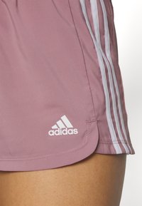 Pantalones cortos Adidas de color rosa claro con diseño de tres rayas blancas a lo largo de los lados y un logo blanco en el dobladillo. Tejido liso y ligero.