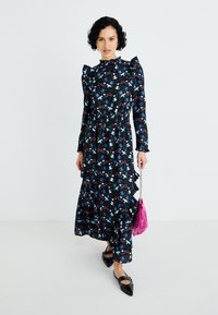 Vestido maxi preto com flores, ombros franzidos, cintura ajustada e saia com camadas. Combinado com uma mala rosa com franjas e sapatos pretos rasos.