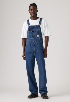Homme portant une salopette en jean bleu avec un t-shirt blanc et des chaussures noires, debout devant un fond clair uni.