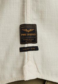 Weißes Stoffetikett mit "PME LEGEND" in schwarzer und goldener Schrift, versehen mit Flügelgrafik. Enthält Pflegeinformationen in schwarzem Stick. 