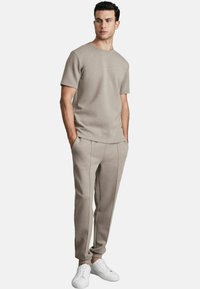 Tricou bej cu mânecă scurtă și guler rotund, asortat cu pantaloni care au o croială slim și dungi pe lateral, purtat cu adidași albi.