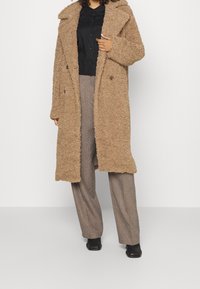 Donna che indossa un lungo cappotto teddy color sabbia sopra una camicia nera con bottoni, pantaloni marroni a gamba larga e stivaletti neri su uno sfondo semplice.