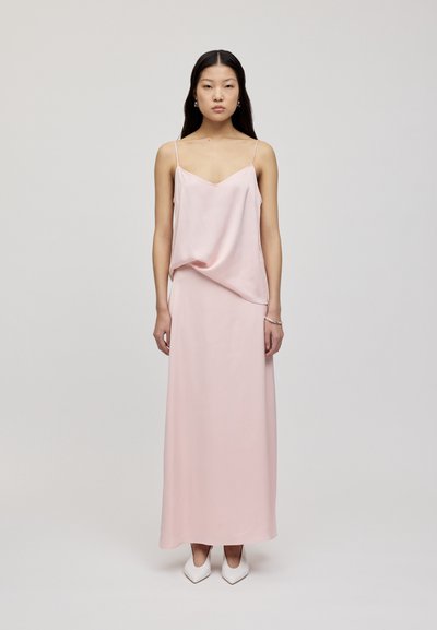 Abito maxi in satin rosa con sottili spalline a spaghetti, una scollatura drappeggiata e una texture liscia. Abbinato a tacchi bianchi a punta.
