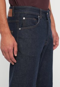 Jeans din denim de culoare bleu închis cu cusături portocalii, buzunare laterale și o etichetă din piele la talie. Prim-plan al ajustării în jurul șoldului.