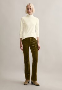 Weißes Langarm-Turtleneck, kombiniert mit olivgrünen Cordhosen. Die Hose verfügt über fünf Taschen und einen Reißverschluss, ergänzt durch beige Stiefel mit Absatz.
