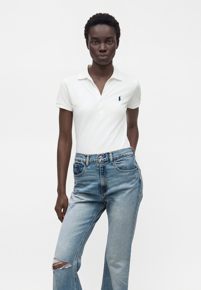 Femme aux cheveux courts et bouclés portant un polo blanc et un jean bleu clair déchiré, debout devant un fond blanc uni.