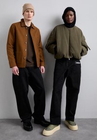 Dos personas están de pie una al lado de la otra. Una lleva una chaqueta marrón con botones, una camisa marrón y pantalones negros. La otra tiene una chaqueta bomber verde oliva, pantalones negros y zapatos verdes.