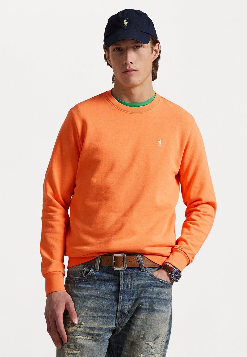 Orange cotton sweatshirt with crew neckline and embroidered logo, paired with blue distressed jeans and leather belt, accessorized with a navy cap.