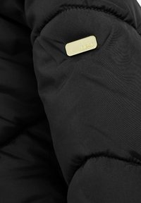 Manche de doudoune noire avec texture matelassée et une petite étiquette en métal doré inscrite "next", mettant en avant un design élégant et discret.