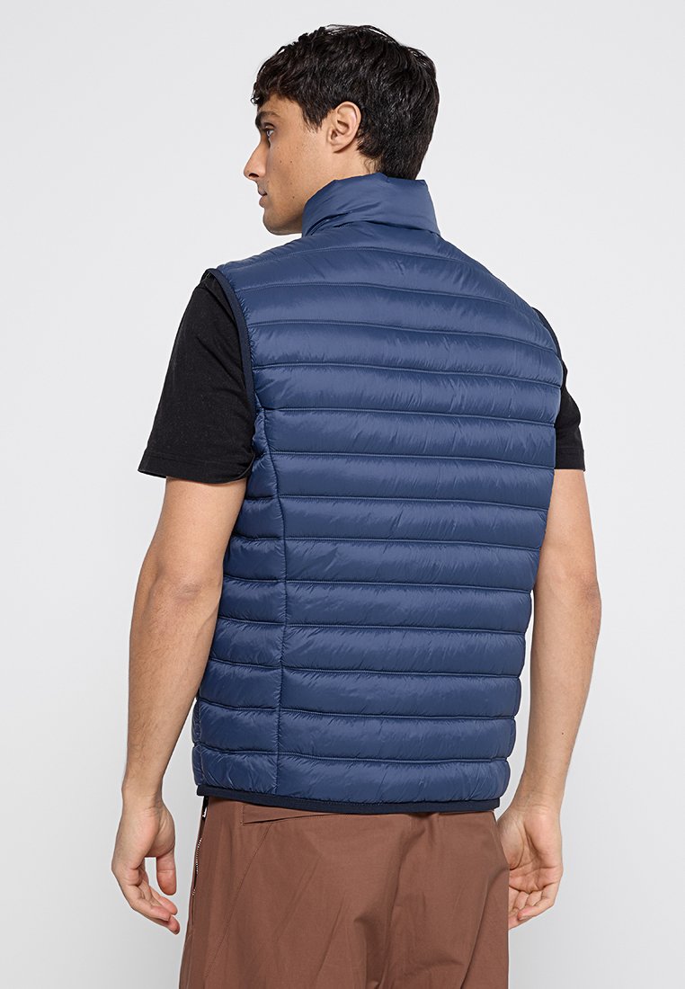 Granatowa kamizelka puchowa z wysokim kołnierzem, z pikowanymi poziomymi panelami i miękką, gładką teksturą. Noszona na czarnej koszuli.