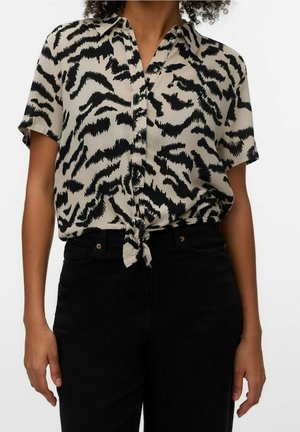 Chemise beige à manches courtes avec imprimé abstrait noir nouée à la taille, portée avec un pantalon noir taille haute sur une personne debout.