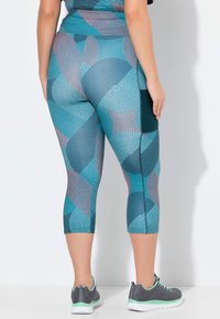 Strakke capri legging in teal met een geometrisch patroon van verschillende vormen in donkerblauw en koraal. Inclusief een zijzak.