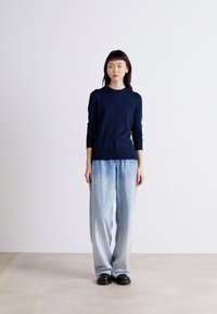 Ναυτικό πλεκτό πουλόβερ με στρογγυλή λαιμόκοψη και ριχούς στριφώματος, συνδυασμένο με ανοιχτό μπλε ξεθωριασμένα παντελόνια wide-leg και μαύρα παπούτσια.