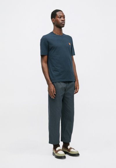 Tricou din bumbac bleu închis cu un mic logo portocaliu pe piept, asociat cu pantaloni largi gri închis și mocasini bicolori.