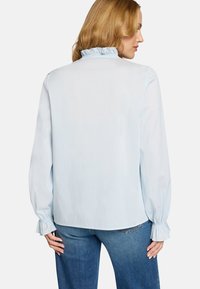 Blouse bleu clair en tissu lisse. Présente un col et des poignets à volants, avec des manches longues et une coupe décontractée. Portée avec un jean en denim.