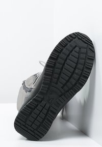 Zapatilla deportiva gris con una suela de goma texturizada y un patrón de banda de rodadura profundo, parte superior sintética con detalles de malla y cordones en contraste.