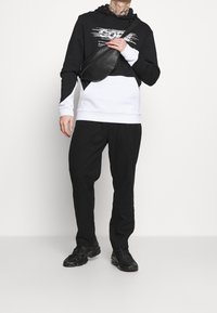 Černá a bílá mikina s kapucí s grafickým designem, spojená s černými kalhotami a teniskami. Obsahuje černou přes rameno tašku.