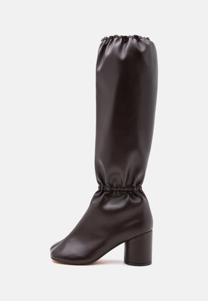 Botte haute marron foncé avec tige froncée et talon bloc sur un fond uni clair.