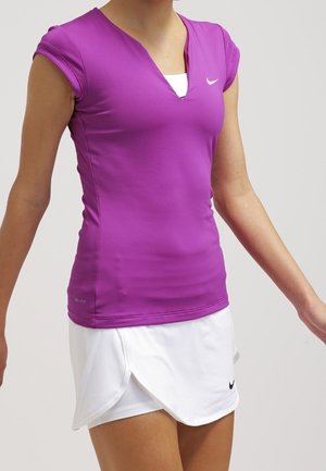 Person iført en tætsiddende lilla kortærmet top og et hvidt sportsskjørt med Nike-logoer, står med armene let udstrakt.