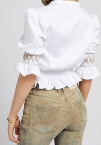 Hvid top med pufærmer, smocket talje og volangkant; har intrikate brune broderier på ærmerne. Båret med falmede, beige jeans.