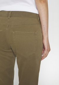 Personne portant un pantalon kaki et un haut blanc, montrant une poche arrière et l'avant-bras avec un petit tatouage vertical en pointillés.