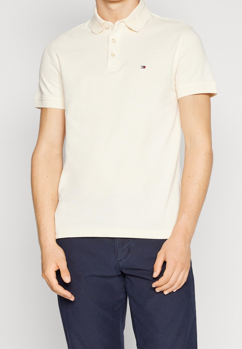 Homme portant un polo crème à manches courtes avec un petit logo sur la poitrine et un pantalon bleu marine foncé, debout devant un fond uni.