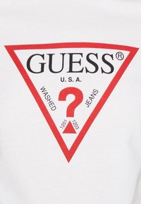 T-shirt bianca con un grande logo a forma di triangolo rosso che presenta il testo "GUESS" e "WASHED JEANS", un punto interrogativo e i numeri 1201, 1203.