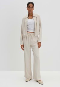 Beige leichte Jacke mit Kragen, kombiniert mit hoch taillierten, weit geschnittenen Hosen und einem weißen, figurbetonten Oberteil. Weiche Textur, minimalistisches Design, lässige Passform.